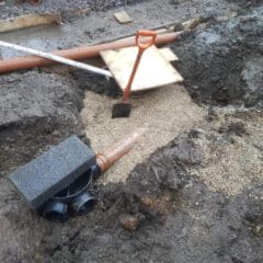 A muddy construction site with exposed pipes, gravel, and a shovel. A T-junction pipe is visible near the bottom, with two black pipe openings and a smaller connected pipe extending out from the gravel-covered trench.