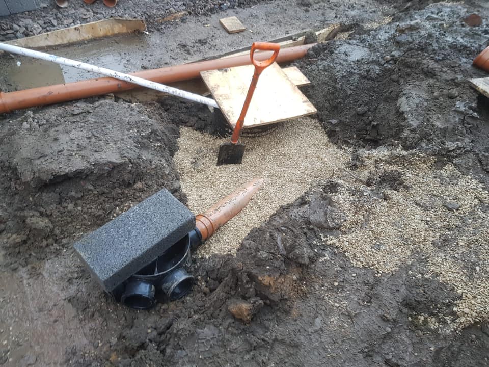A muddy construction site with exposed pipes, gravel, and a shovel. A T-junction pipe is visible near the bottom, with two black pipe openings and a smaller connected pipe extending out from the gravel-covered trench.