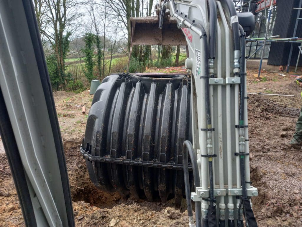 An excavator places a large black cylindrical tank into a hole in a muddy construction site surrounded by trees. Scaffolding is visible on the right side.