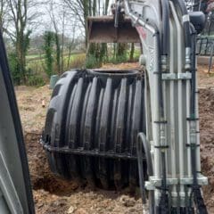 An excavator places a large black cylindrical tank into a hole in a muddy construction site surrounded by trees. Scaffolding is visible on the right side.