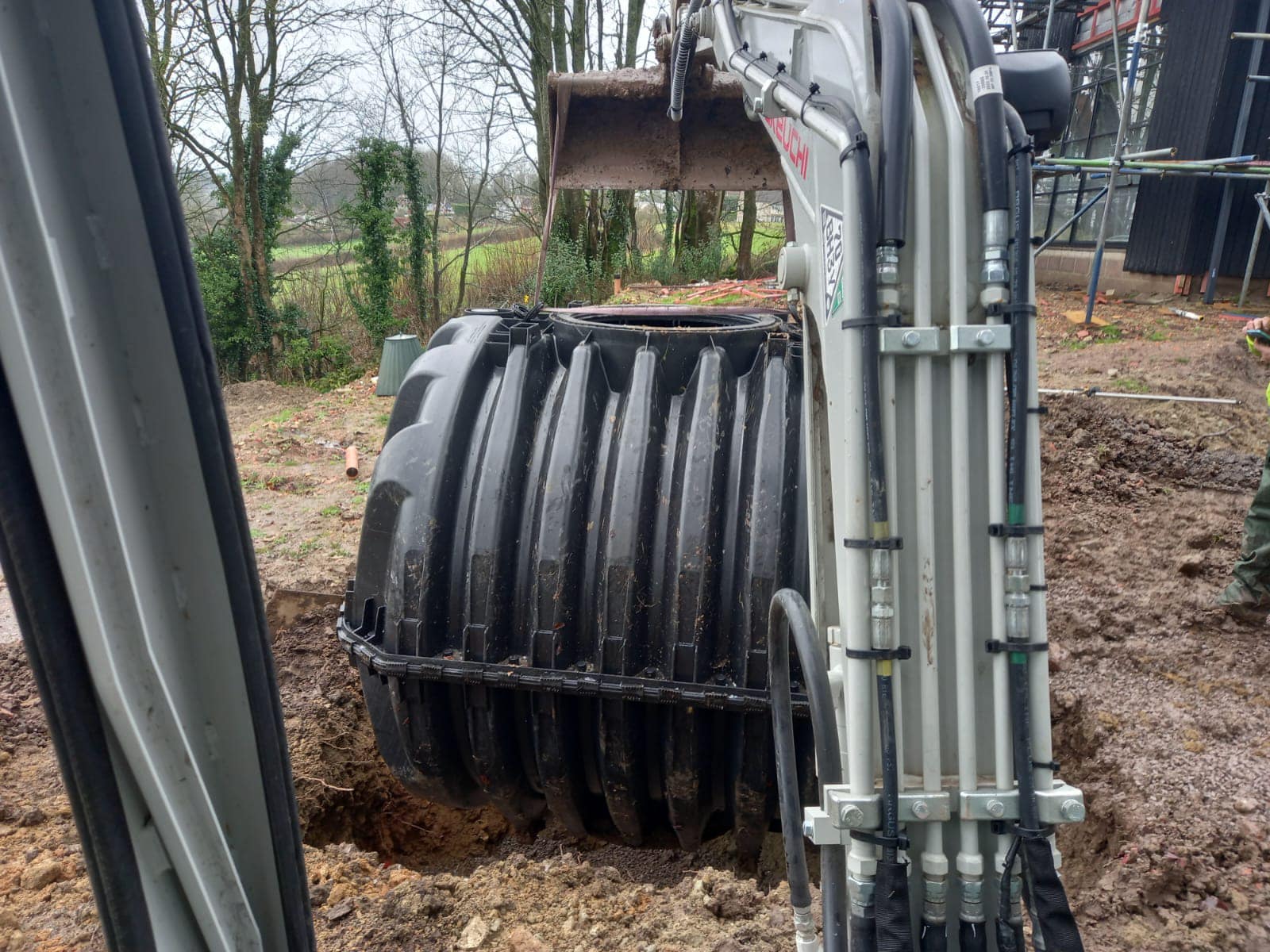 An excavator places a large black cylindrical tank into a hole in a muddy construction site surrounded by trees. Scaffolding is visible on the right side.