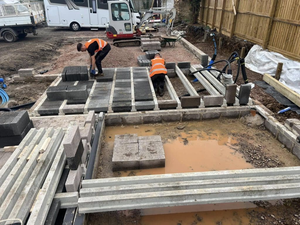 Two workers in orange safety vests are placing large concrete slabs in a construction area. The ground is uneven and muddy, with water pooled in some parts. A motorhome is parked in the background near construction equipment.