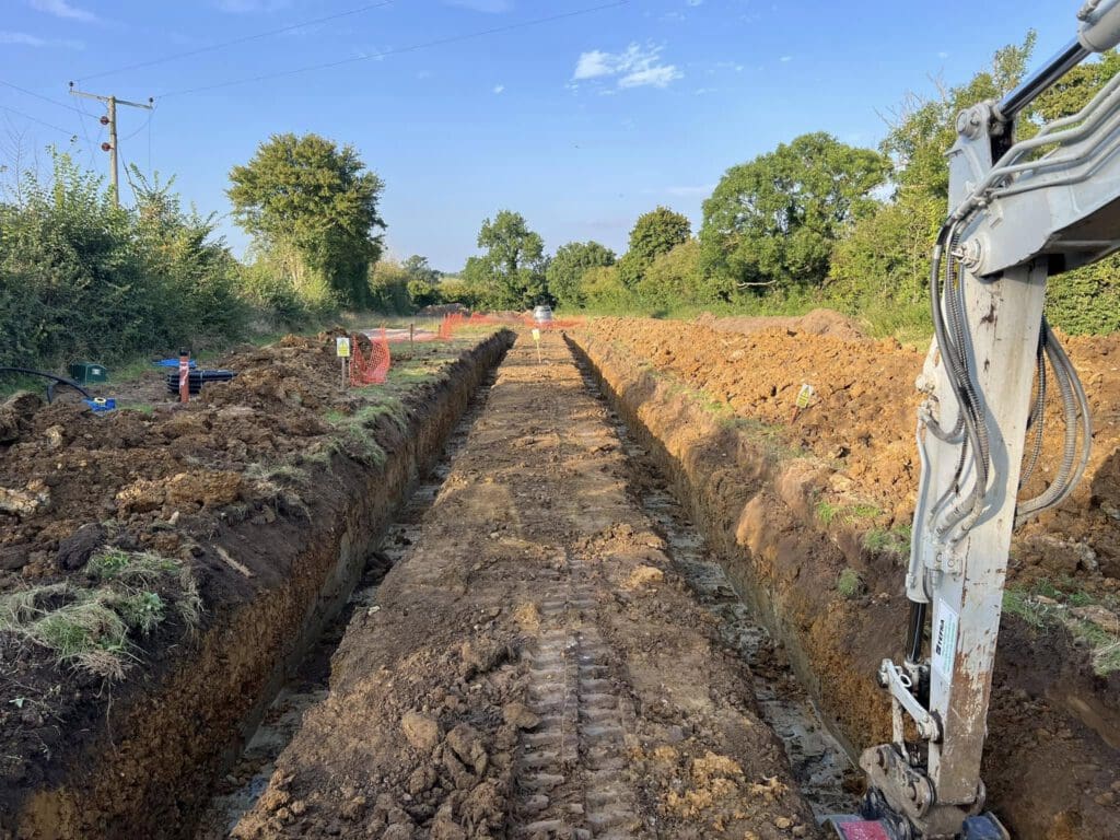 A construction site with a long trench dug into the earth. A small excavator sits on the right side. The area is surrounded by grass and trees under a clear blue sky. Red markers and equipment line the trench.