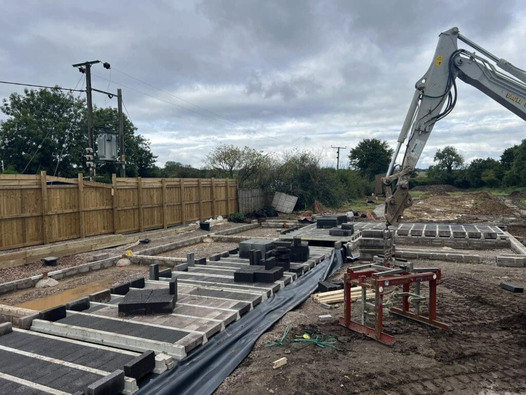 Construction site with a digger arm on the right, laying concrete blocks on the ground. A wooden fence and trees line the left side. Overcast sky above. Various tools and materials scattered around the area.