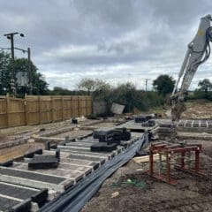 Construction site with a digger arm on the right, laying concrete blocks on the ground. A wooden fence and trees line the left side. Overcast sky above. Various tools and materials scattered around the area.