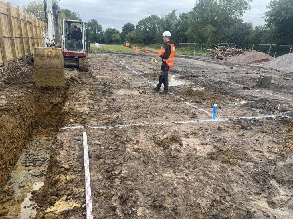Construction site with muddy ground, where a worker in an orange vest and hard hat stands near a mini excavator digging a trench. Measuring tapes and marked lines are visible. Trees and a wooden fence line the perimeter. Overcast sky above.