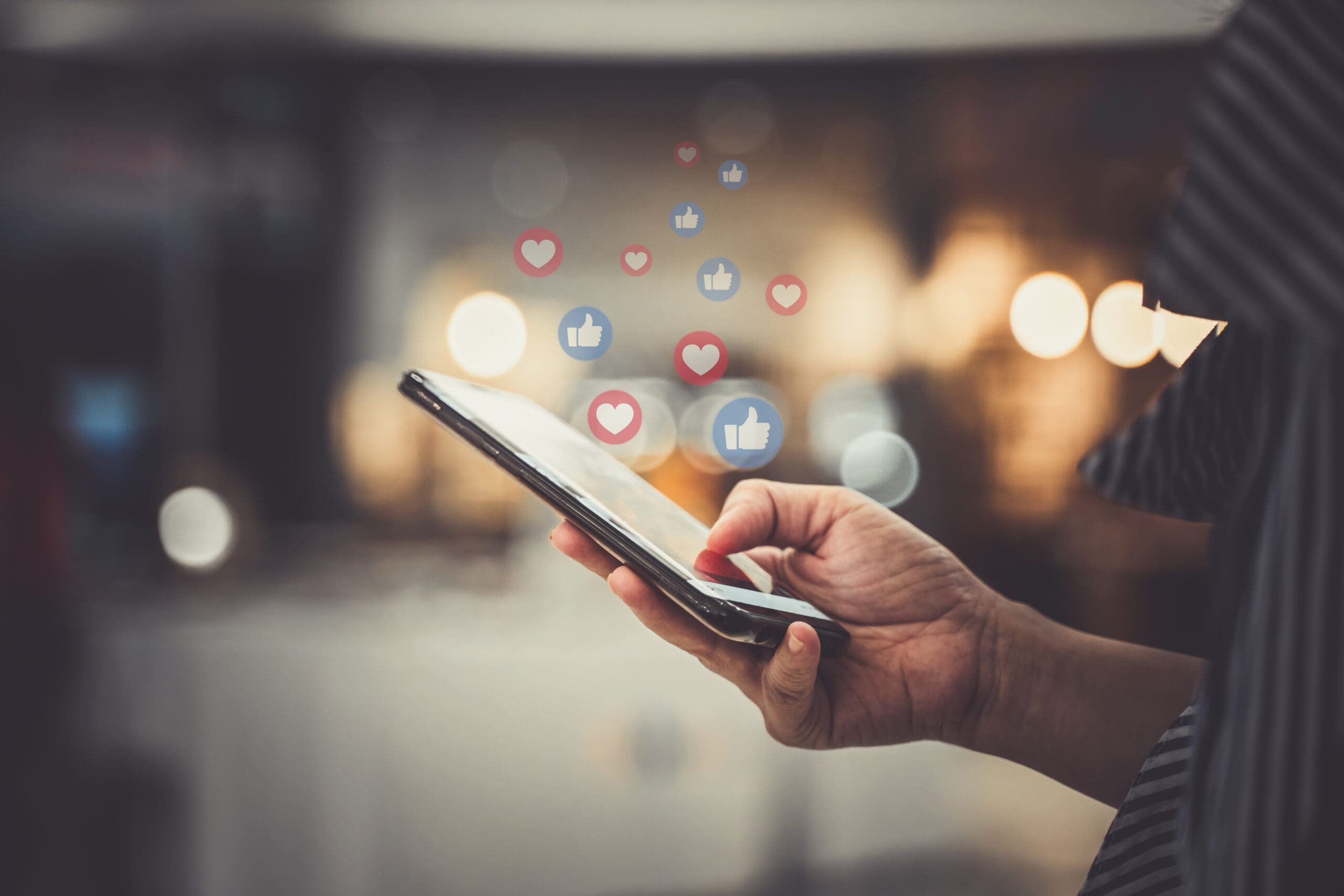 A person holding a smartphone with floating social media icons above the screen, including hearts and thumbs-up symbols. The background is blurred with warm lighting, creating a cozy atmosphere.