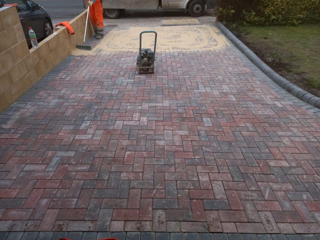 A nearly completed driveway with red and gray interlocking bricks in a herringbone pattern. A compacting machine sits on the bricks. Two workers are seen in the background near a wall. A truck is parked on the road.
