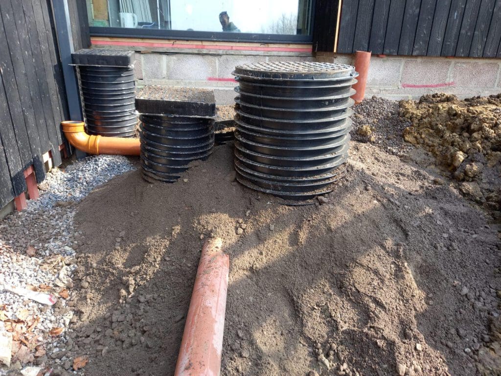 Plastic drainage pipes and covers stacked beside a building exterior with a window. Piles of sand and gravel surround the area, accompanied by exposed orange pipes. A portion of the soil is recently disturbed, indicating ongoing construction work.