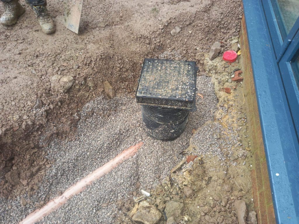 A partially covered drainage pipe is surrounded by dirt and gravel next to a brick wall. There's a black plastic cover on top of the pipe. A red cap and a person's muddy boots are visible in the background.