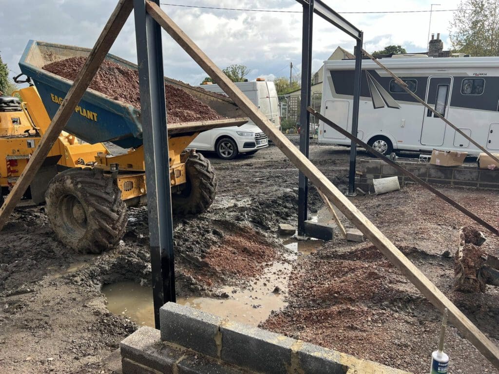 Construction site with a yellow dump truck unloading gravel onto muddy ground. Steel frame structures are partially erected nearby. A white car and a caravan are parked in the background on a cloudy day.