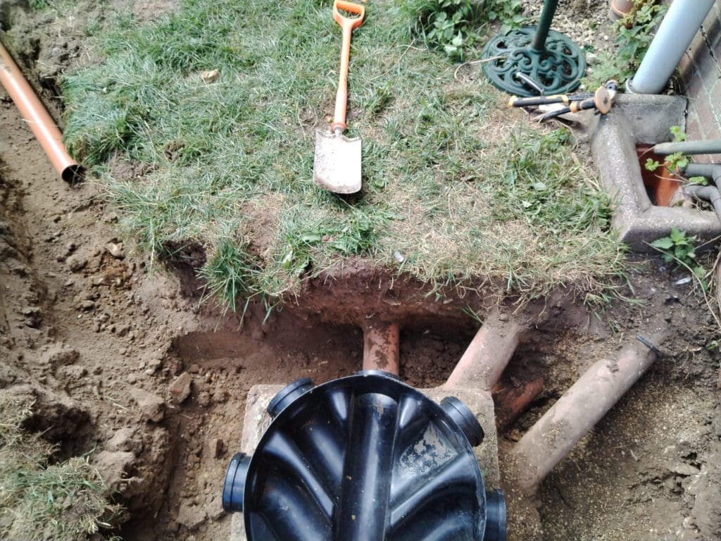 A garden area with a dug-up section showing exposed pipes and a black drainage junction. A small shovel with an orange handle is resting on the grass. Various tools are scattered around the edge of the excavation site.