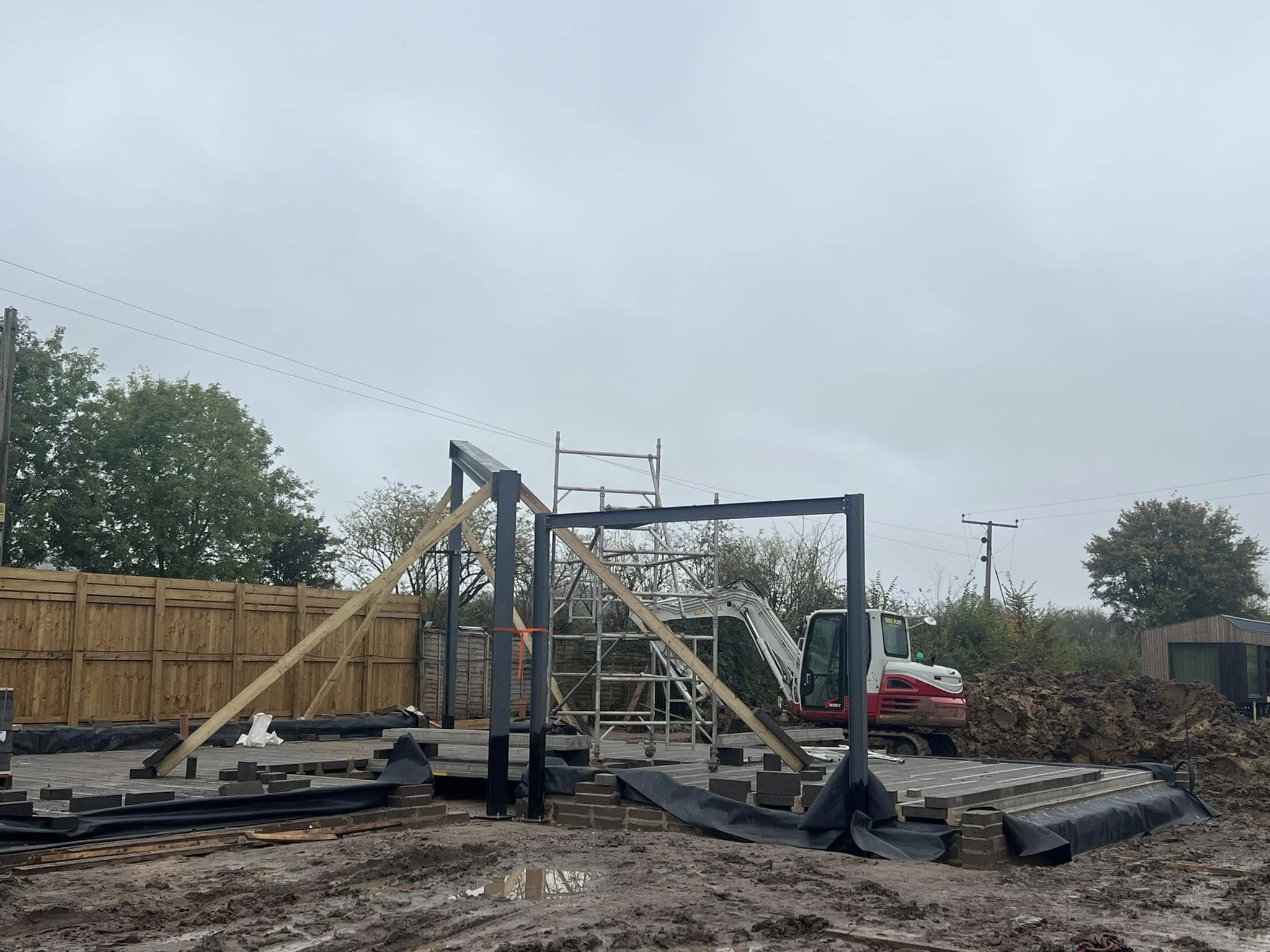 Construction site with a partially built steel frame structure. Wooden beams support the frame. A small excavator is nearby, and scaffolding is set up. The ground is muddy, and the sky is overcast. A wooden fence and trees are in the background.