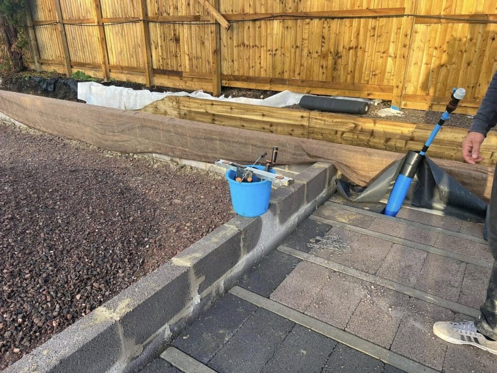 A garden area under construction. There are concrete blocks forming a low wall, a blue bucket with tools on top, and landscaping fabric covering soil. A wooden fence runs along the background, and a person's arm is visible on the right.