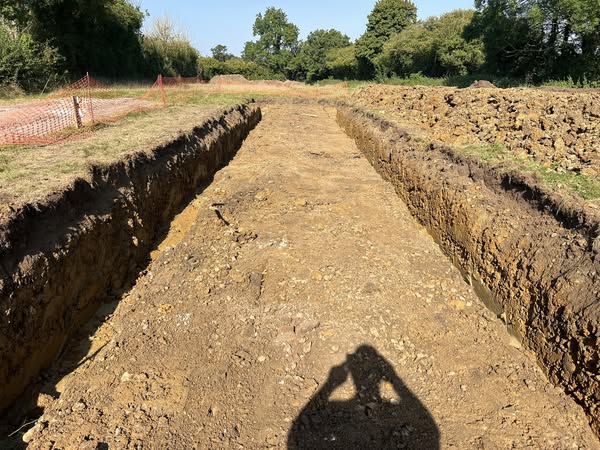 A long, narrow trench dug into the earth, lined by mounds of dirt on both sides. Green trees and grass surround the area. The shadow of a person taking a picture is visible in the foreground.