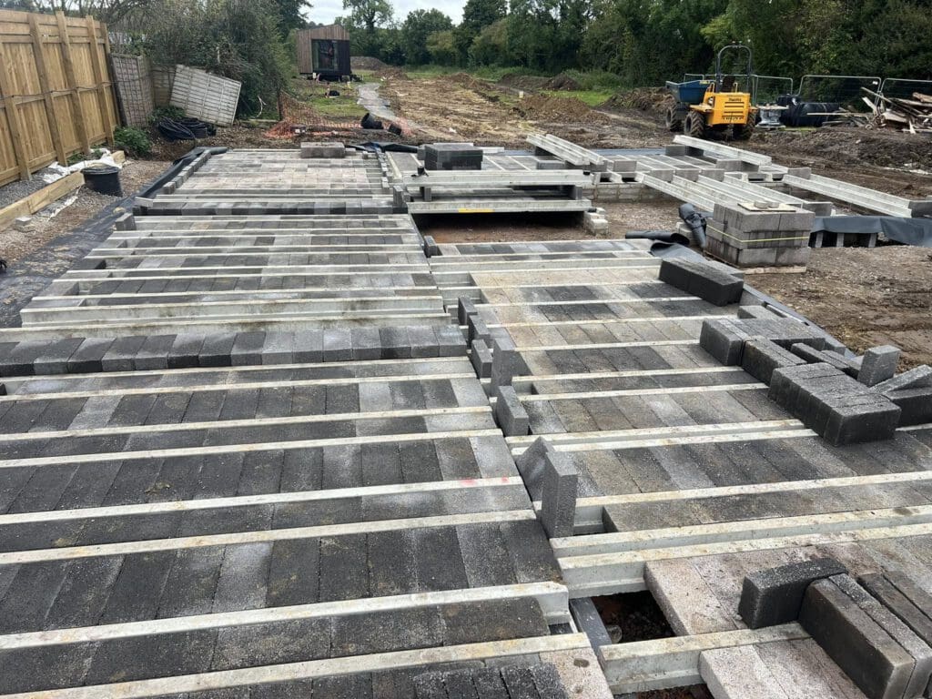 Construction site with concrete slabs and steel beams laid on the ground. There are various building materials and a small yellow digger in the background, with trees and a wooden fence along the perimeter.
