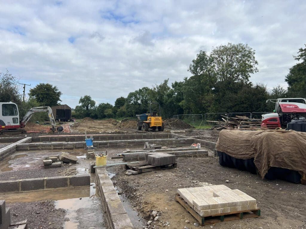 Construction site with unfinished building foundations surrounded by blocks and construction materials. A small excavator and a yellow compact loader are visible. Trees and partly cloudy sky in the background.