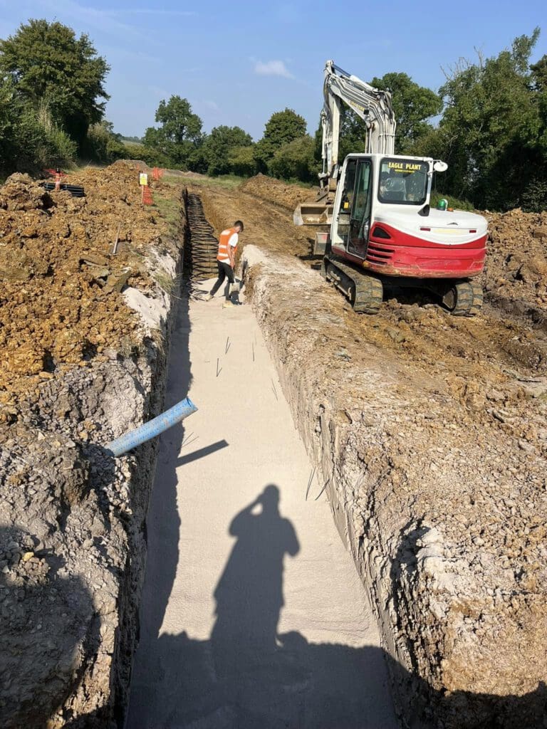 A construction site with a deep trench dug in the ground. A worker in a high-visibility vest stands examining the trench. A small excavator is parked nearby. Trees and dirt piles are in the background, and a shadow of a person is visible in the foreground.