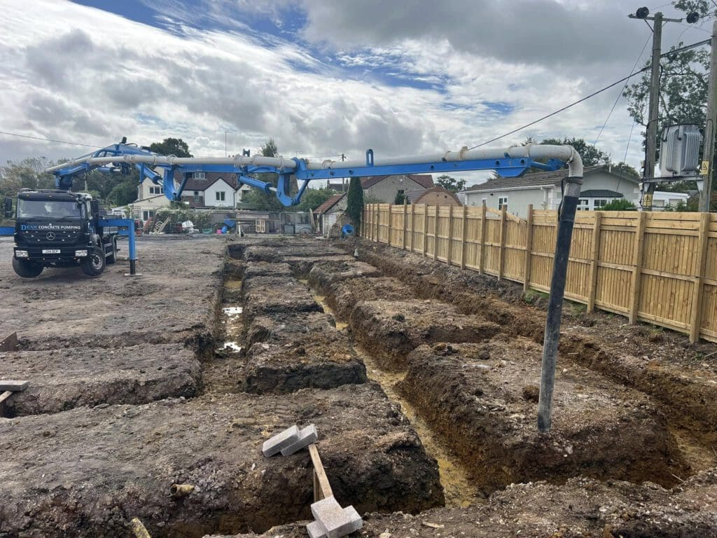 A construction site featuring deep trenches in the ground, partially filled with concrete. A blue concrete pump truck is positioned on the left, extending its arm to pour concrete. A wooden fence lines the right side, with trees and houses in the background.