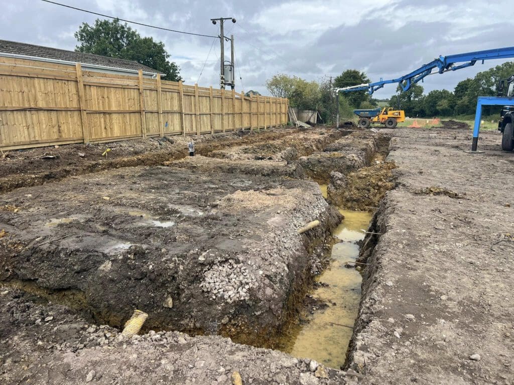 Construction site with trenches dug in the soil, muddy water pooled at the bottom. A wooden fence lines the left side. A blue crane and yellow excavator are visible in the background under a cloudy sky.