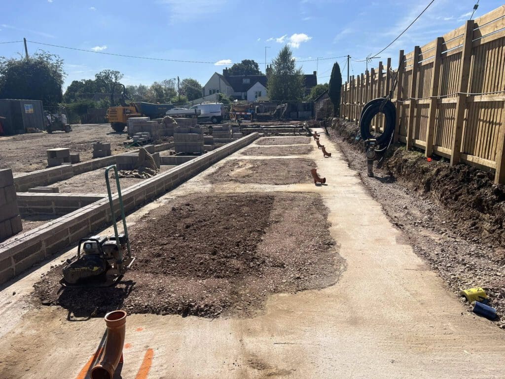 A construction site with leveled ground and partially built walls. Piles of earth and construction materials, including blocks and pipes, are visible. A wooden fence runs along the right side, with trees and a house in the background.