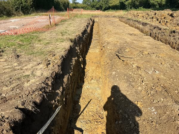 A trench stretches through an open field under sunny skies. Freshly dug soil lines the trench edges. An orange safety net is visible in the background. A person's shadow appears on the ground near the trench.