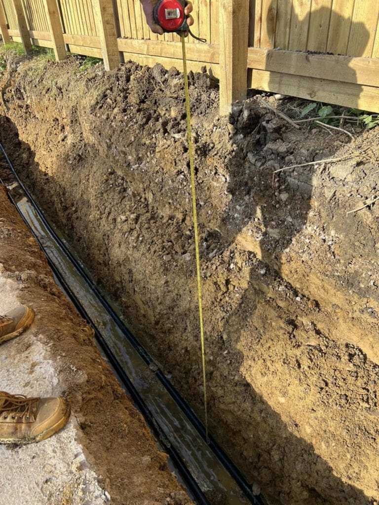 A deep trench near a wooden fence is being measured with a yellow tape measure. The trench contains black plastic pipes and is surrounded by dirt. A person's hand holds the tape measure, and brown work boots are visible at the edge.