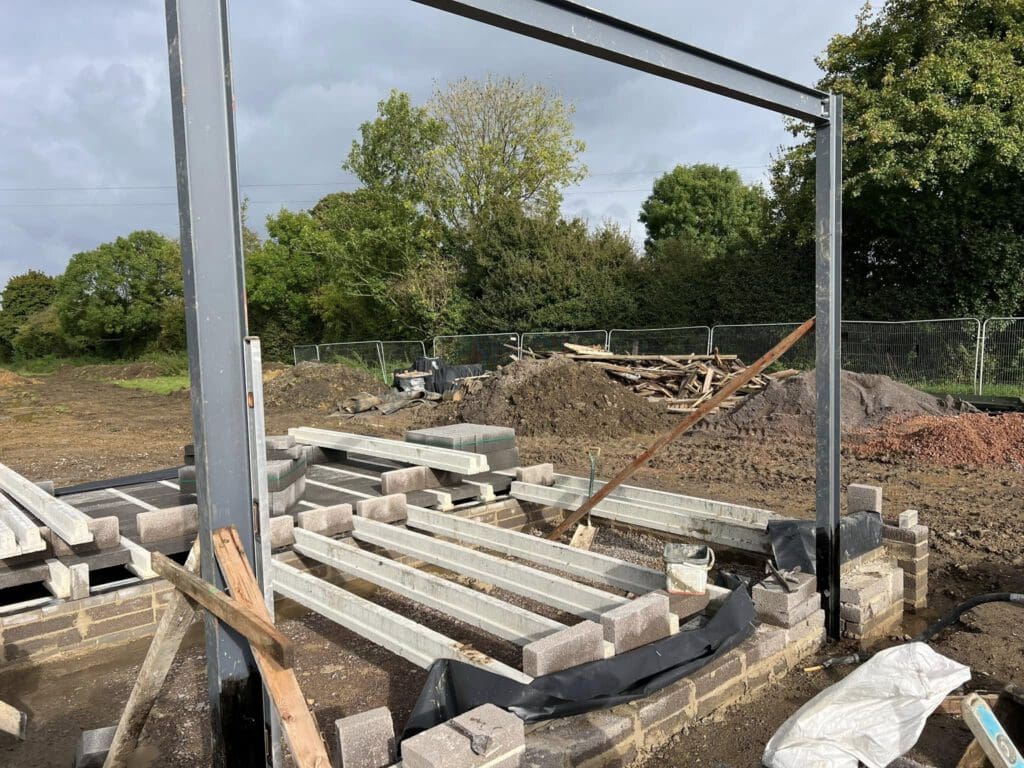 Construction site with metal beams and concrete blocks arranged on the ground. A partially built wall is visible, surrounded by piles of soil and wooden planks. Trees and fencing are in the background under a cloudy sky.