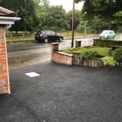 A driveway with newly laid asphalt is in the foreground. A brick wall borders the driveway, and a small garden area is visible. Two cars are parked on the wet street beyond the driveway, surrounded by green trees and grass.