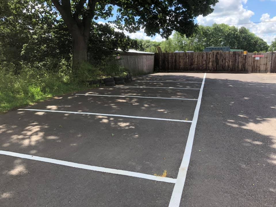 An empty outdoor parking lot with clearly marked parking spaces. A large tree provides shade on the left side. There's a wooden fence and greenery in the background, under a partly cloudy sky.